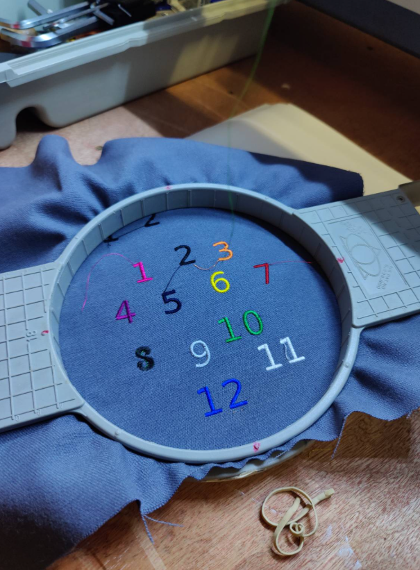 A grey hoop with grey fabric mounted in it. the numbers 1 to 12 are stitched out in different colours. Apart from 8 which appears to be mangled.