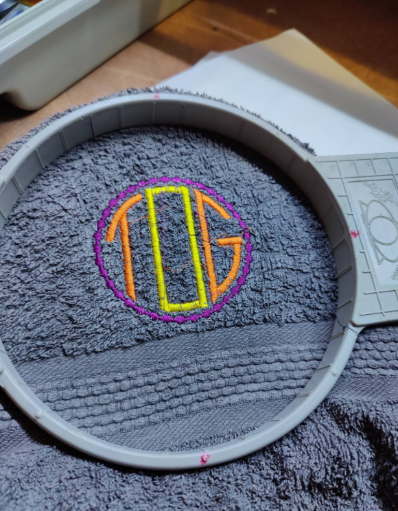 A finished monogram in the hoop. IT's a grey towel with Tog in orange and yellow letters in a purple circle.