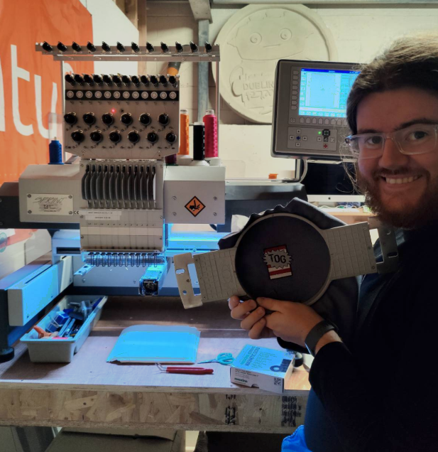 A tog member stands in front of the embroidery machine, showing off a piece of embroidery in a hoop