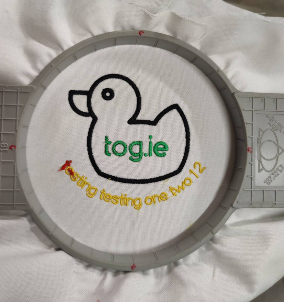 An early test embroider. The white fabric is still mounted in the hoop. It has a black duck with the text tog.ie in green. Below that is testing testing one two 