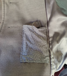 A grey patch pocket attached to the inside of a grey coat. A battery pack and wire are poking out of the pocket.
